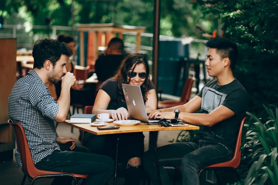 Three young people sitting at a table at outdoor restaurant, one has Apple laptop open.